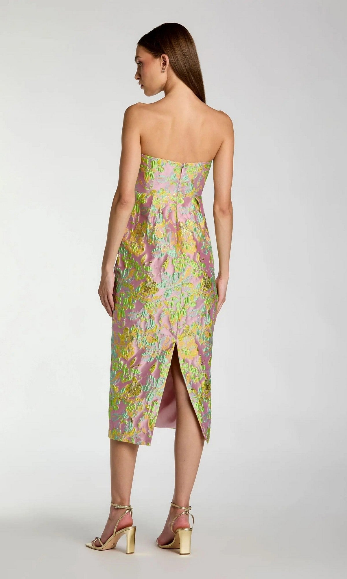 Wearing the Mac Duggal 56423 Midi Wedding Guest Dress—a strapless brocade design featuring a pastel floral pattern and back slit—this woman completes the elegant look with gold high-heeled sandals, standing against a plain background.